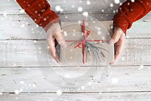 A woman in a red sweater holding a Christmas gift with a red ribbon and snow over on a wooden table.