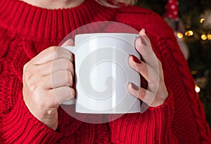 Woman in red sweater with white mug in hands against Christmas tree with lights, mock up