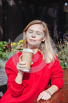 Woman in a red sweatshirt sitting in a cafe and holding a disposable cup of coffee.