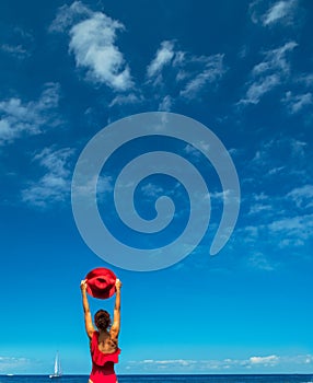 Woman in red swimming suit holds red hat in her hands. Calm ocean water and pure blue sky at the background