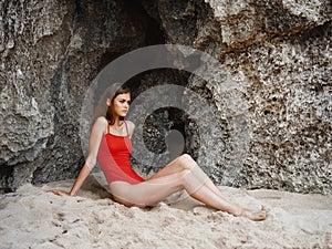 Woman in red swimsuit lying by the rocks on the sand looking at the camera with a beautiful tan from the sun on the