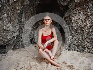 Woman in red swimsuit sitting by the rocks on the sand with a beautiful tan from the sun on the beach, travel to Bali, a