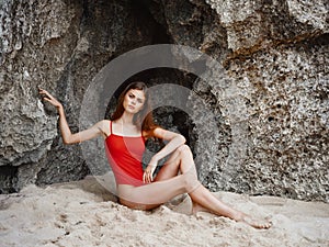 Woman in red swimsuit sitting by the rocks on the sand with a beautiful tan from the sun on the beach, a trip to the sea