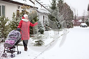 Woman in red warm long jacket stands on small street at winter season, gesturing by hand on snowy road while walking with baby