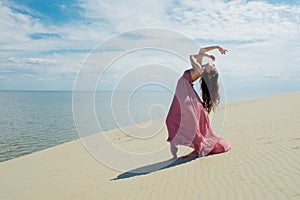 Woman in red waving dress with flying fabric runs on background of dunes. Gymnast on the back of the dune