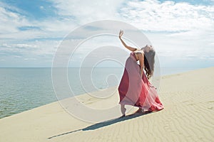 Woman in red waving dress with flying fabric runs on background of dunes. Gymnast on the back of the dune