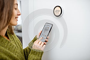 Woman regulating heating temperature with phone and thermostat at home