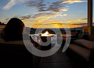 A woman relaxes with a glass of wine at night in front of an outdoor firepit on a patio of a luxury home overlooking a city.