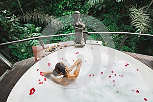 Woman relaxing bath tub full of foam outdoors with jungle view. View from behind