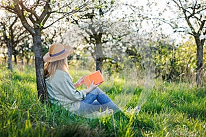 Woman is relaxing in blooming orchard and reading book to improve her mindfulness and mental health