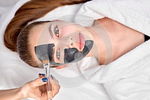 Woman relaxing with charcoal facial mask, cropped professional beautician applying on face