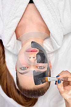 Woman relaxing with charcoal facial mask, cropped professional beautician applying on face