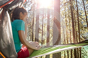 Woman relaxing and drinking in hanging tent camping in forest woods during sunny day.Group of friends people summer