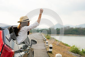 Woman relaxing and enjoying road trip. Young Asian happines female wearing hat inside compact black car with raise her hand out to