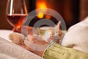 Woman relaxing by the fire, holding a glass of wine and her cute ginger kitten - close up on hands