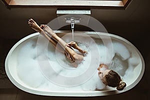 Woman relaxing in foam bath with bubbles in dark bathroom by window