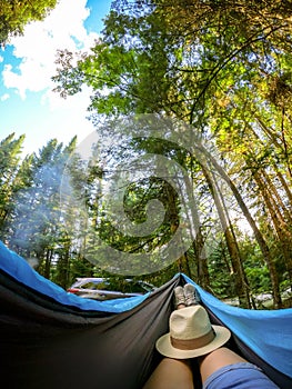 Woman relaxing in the hammock by the lake in the forest, POV view of legs in trekking boots with straw hat. Wanderlust concept