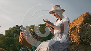 Woman is relaxing in meadow with smartphone in hands at sunset. Young girl holds mobile phone in hands while resting in park. Fema