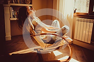 Woman relaxing and napping in comfortable modern chair near window radiator, livingroom. Warm natural light. Cozy home. Casual clo