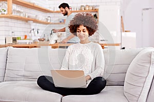 Woman Relaxing Sitting On Sofa At Home Using Laptop Computer With Man In Kitchen Pouring Drink