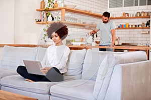 Woman Relaxing Sitting On Sofa At Home Using Laptop Computer With Man In Kitchen Pouring Drink