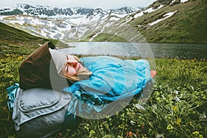 Woman relaxing in sleeping bag laying on grass enjoying lake and mountains