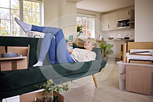 Woman Relaxing On Sofa Using Mobile Phone Surrounded By Unpacking Boxes After Moving Into New Home