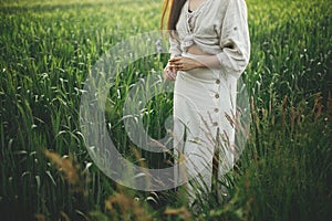 Woman relaxing in wheat field in evening, cropped view. Stylish young female in rustic dress holding wildflowers in hands in