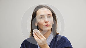 Woman removing makeup from face. Female in blue bathrobe removes cosmetics from face and looking in mirror. Makeup removing. Healt