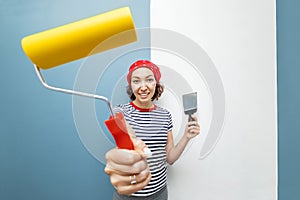 Woman repairman poses with a roller and trowel against the background of pasted wallpaper and a plastered wall