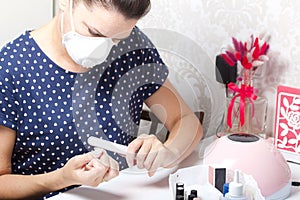 A woman with a respirator on her face gets a manicure. Removes old nail Polish with a nail buff. Next to a set of lacquers, tools