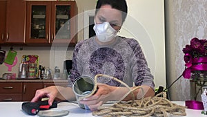 A woman in a respirator wraps a tourniquet around a tin can. Fastens the tourniquet with a glue gun. Self-isolation crafts