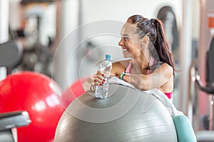 Woman resting on a fitness ball in the gym holds water bottle and laughs looking aside