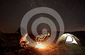 Woman resting at night camping near campfire, tourist tent, bicycle under evening sky full of stars