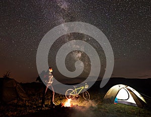 Woman resting at night camping near campfire, tourist tent, bicycle under evening sky full of stars