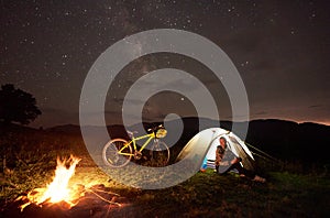 Woman resting at night camping near campfire, tourist tent, bicycle under evening sky full of stars