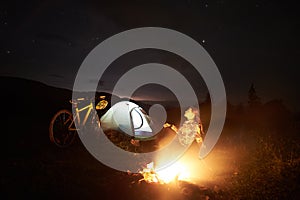 Woman resting at night camping near campfire, tourist tent, bicycle under evening sky full of stars