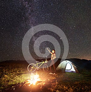 Woman resting at night camping near campfire, tourist tent, bicycle under evening sky full of stars