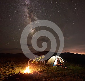 Woman resting at night camping near campfire, tourist tent, bicycle under evening sky full of stars