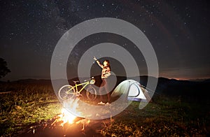 Woman resting at night camping near campfire, tourist tent, bicycle under evening sky full of stars