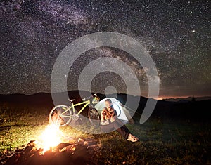 Woman resting at night camping near campfire, tourist tent, bicycle under evening sky full of stars