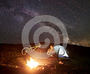 Woman resting at night camping near campfire, tourist tent, bicycle under evening sky full of stars