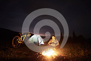 Woman resting at night camping near campfire, tourist tent, bicycle under evening sky full of stars