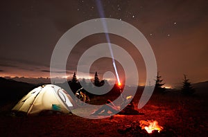 Woman resting at night camping near campfire, tourist tent, bicycle under evening sky full of stars