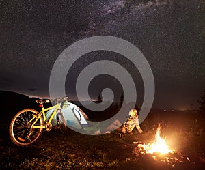 Woman resting at night camping near campfire, tourist tent, bicycle under evening sky full of stars