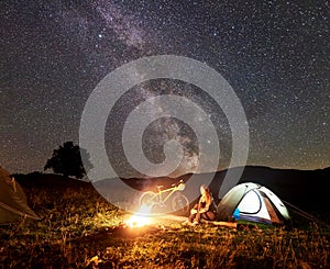 Woman resting at night camping near campfire, tourist tent, bicycle under evening sky full of stars