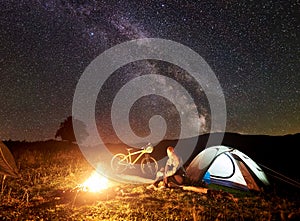 Woman resting at night camping near campfire, tourist tent, bicycle under evening sky full of stars