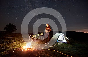 Woman resting at night camping near campfire, tourist tent, bicycle under evening sky full of stars