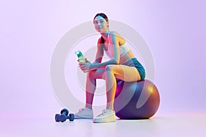 Woman resting between trainings sitting on fit-ball with battle of water in neon light against gradient studio photo