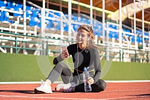 Woman resting after workout or running, sitting on a treadmill rubber stadium, using and listens to music with wired headphones on